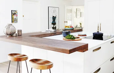 Modern kitchen with a wooden countertop, stools, a cooktop, and a blue pot. A poster and decor items are visible.