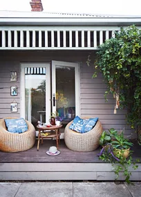 Front porch with two rattan chairs, blue pillows, table with coffee, and climbing plants. Cozy outdoor space.