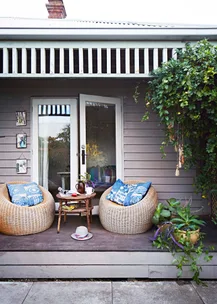Front porch with two rattan chairs, blue pillows, table with coffee, and climbing plants. Cozy outdoor space.