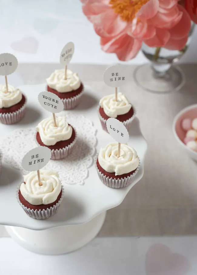 Valentine cupcakes with white frosting, topped with mini signs saying "Be Mine," "You're Cute," and "Love."