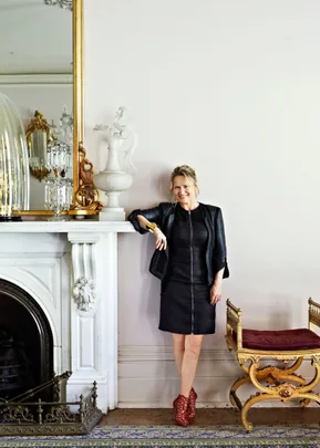 A woman in a black dress and red shoes leans on a white fireplace in an elegant room with gold accents.