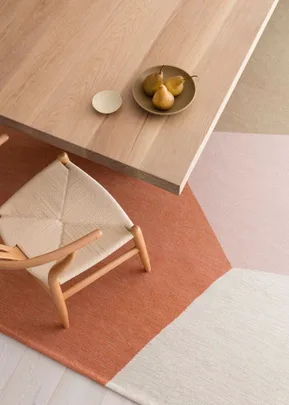 A wooden chair beside a table with a plate of pears; displayed on a geometric patterned rug in earthy tones.