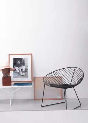 Modern room with a black wire chair, framed photo, books, and a sculpture on a white table against a plain wall.