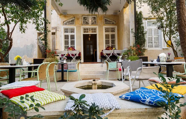 Outdoor cafe with colorful cushions, tables, and chairs in a cozy courtyard setting with plants and potted flowers.