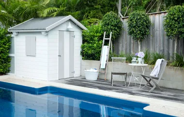 Poolside scene with a white shed, two chairs, a table with flowers, and a towel on a ladder, surrounded by greenery.