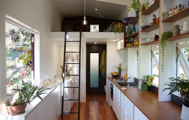 Interior of a compact modern kitchen with wooden countertops, plants, a ladder to a loft, and natural light from windows.
