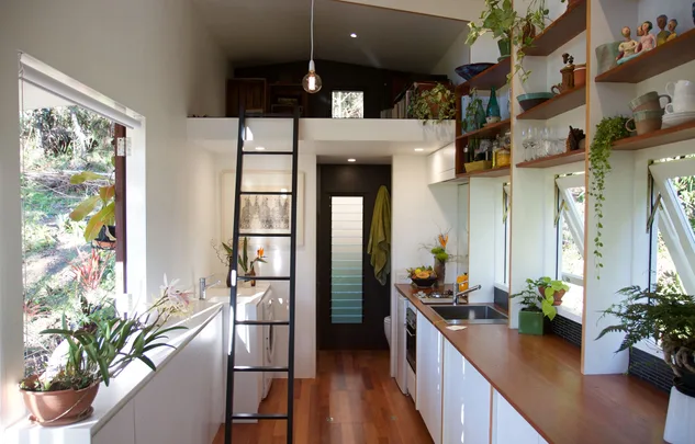 Interior of a compact modern kitchen with wooden countertops, plants, a ladder to a loft, and natural light from windows.