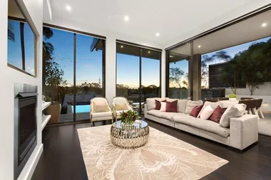 Modern living room with large windows, beige sofa, pink cushions, patterned rug, and scenic views of trees and sunset.