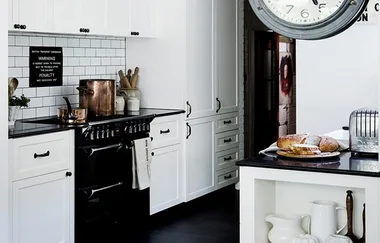 "White kitchen with black countertops, wall clock, stove, and bread on a plate. Copper kettles and jars on counter."