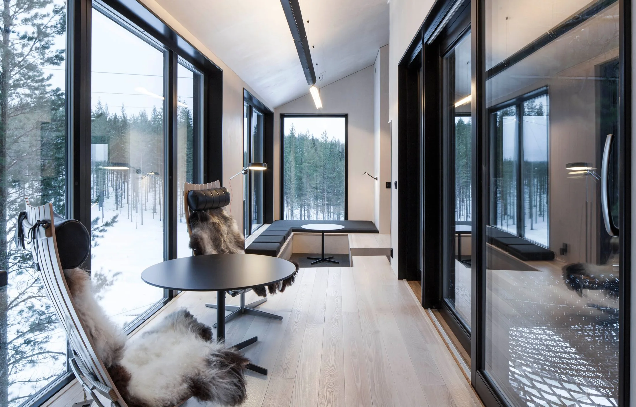 Interior of modern cabin with large windows, forest view, sleek furniture, and fur-covered chairs.