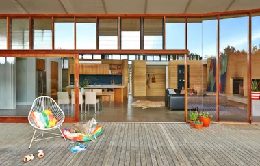 Outdoor patio with a chair, colorful pillows, a dog resting, and a view of the modern interior through large glass doors.