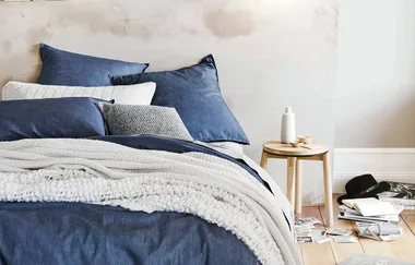 Cozy bed with blue and white pillows, blanket; wooden nightstand with decor, books, and hat on the floor nearby.