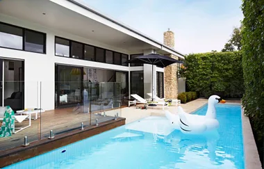 Modern house with pool, large inflatable swan raft, wooden deck, sun loungers, umbrella, glass fence, and greenery.