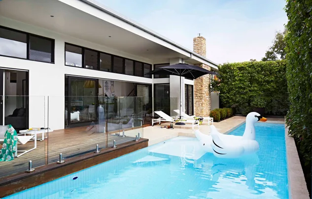 Modern house with pool, large inflatable swan raft, wooden deck, sun loungers, umbrella, glass fence, and greenery.