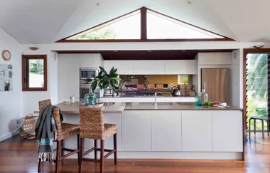 Modern kitchen with white cabinets, stainless steel appliances, island with wicker chairs, and a large triangular window.