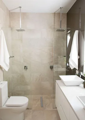 Modern bathroom with dual rain showers, glass walls, toilet, and white basin on vanity cabinet.