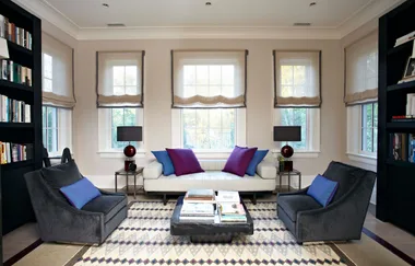 Modern living room with white sofa, colorful pillows, gray chairs, bookshelves, and a patterned rug.