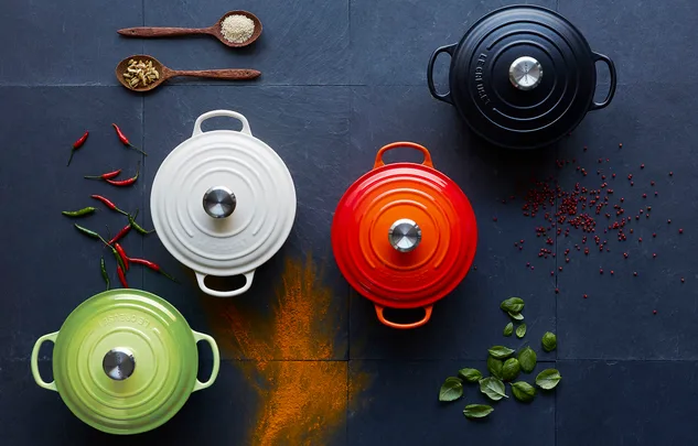 Le Creuset pots in black, white, green, and red on a dark surface with spices, chili peppers, and herbs.