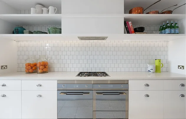Modern white kitchen with gas stove, tiled backsplash, and open shelves displaying dishes and copper pans.