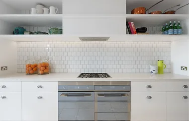 Modern white kitchen with gas stove, tiled backsplash, and open shelves displaying dishes and copper pans.
