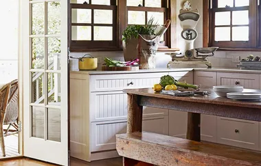 Rustic kitchen with wooden counter, vintage scale, white cabinets, and fresh produce near a sunny window.