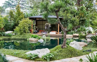 Japanese garden with a pond, stone path, and a gazebo surrounded by lush greenery and trees.