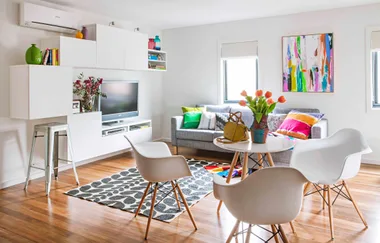 Bright living room with modern furniture, colorful cushions, abstract art, and flowers on a round table.