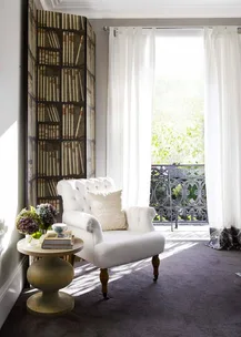 Cozy corner with a white armchair, side table with books and flowers, and a bright window with sheer curtains overlooking greenery.