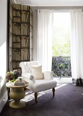 Cozy corner with a white armchair, side table with books and flowers, and a bright window with sheer curtains overlooking greenery.