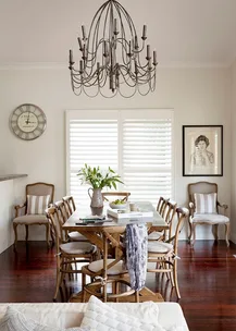 Dining room with wooden table, chairs, chandelier, wall clock, window shutters, and framed black-and-white photo.