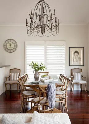 Dining room with wooden table, chairs, chandelier, wall clock, window shutters, and framed black-and-white photo.