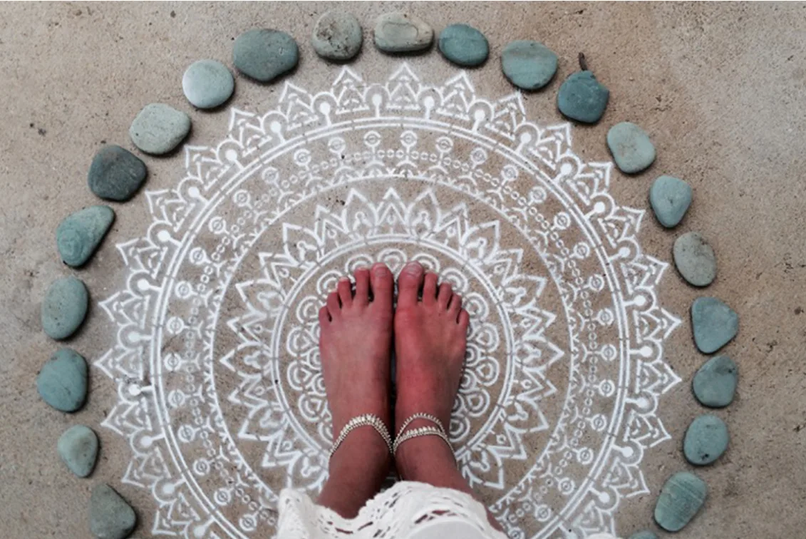 Bare feet with anklets on a mandala drawn on sand, surrounded by smooth stones.
