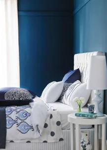 Elegant bedroom with blue walls, white bed, patterned bedding, and a bedside table with a lamp and flowers.
