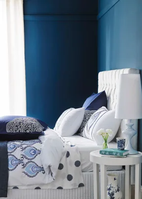 Elegant bedroom with blue walls, white bed, patterned bedding, and a bedside table with a lamp and flowers.