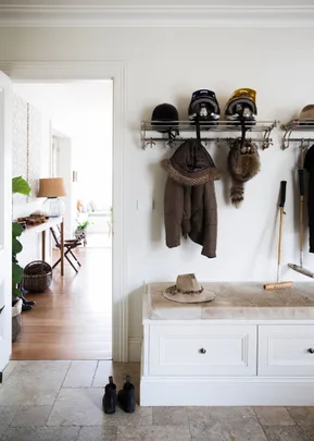 A mudroom with hanging coats, helmets, a fur hat, bench with a hat, boots on the floor, and a walking stick.