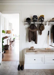 A mudroom with hanging coats, helmets, a fur hat, bench with a hat, boots on the floor, and a walking stick.