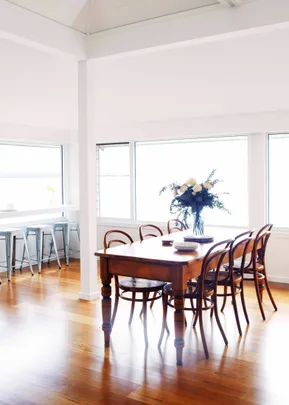 Bright dining room with a wooden table, chairs, and a flower vase. Large windows let in natural light.