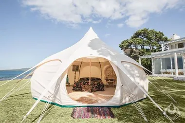 A large, white yurt tent set up on a grassy lawn by the sea, with furniture visible inside under a sunny sky.