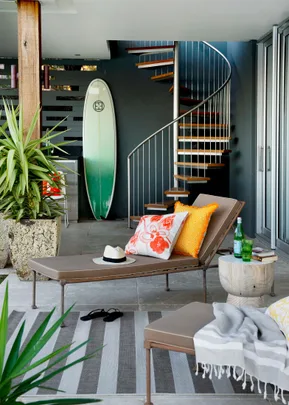 Outdoor lounge with a spiral staircase, surfboard, chaise with colorful pillows, hat, flip-flops, and a small table.
