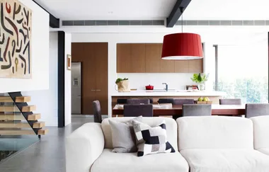 Modern open-plan living area with white couch, dining table, kitchen island, red lamp, and abstract wall art.