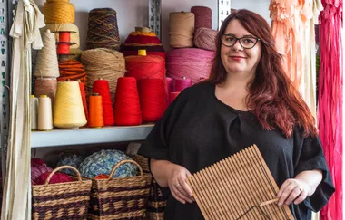 A woman with red hair holds a wooden tool, standing next to shelves filled with colorful spools of yarn.