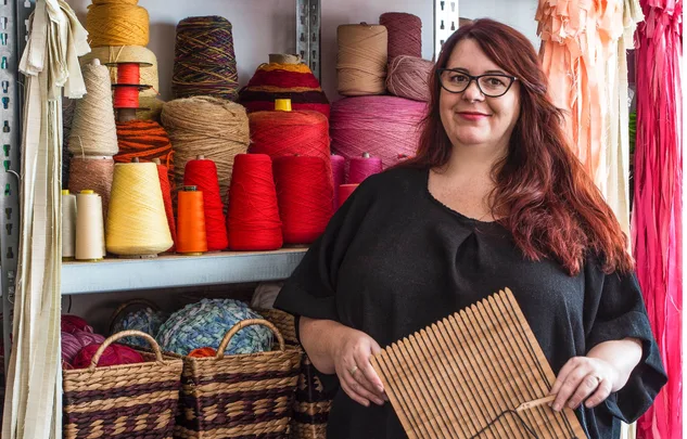 A woman with red hair holds a wooden tool, standing next to shelves filled with colorful spools of yarn.