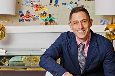 Man in a suit sitting indoors, smiling, with a large beach photograph in the background.