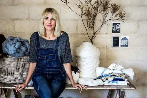 Woman in overalls sitting on a bench, surrounded by wool and textiles, with a dried branch and small photos on the wall.