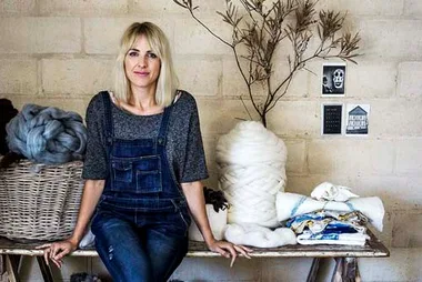 Woman in overalls sitting on a bench, surrounded by wool and textiles, with a dried branch and small photos on the wall.