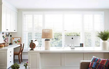 Bright home office with large windows, desk lamp, computer, cushion, and plant.