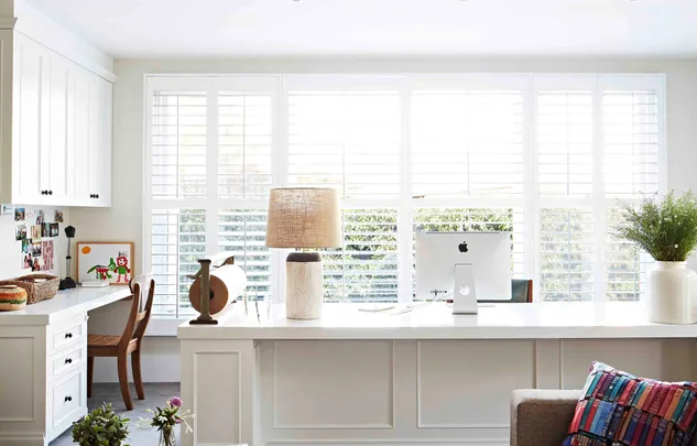 Bright home office with large windows, desk lamp, computer, cushion, and plant.