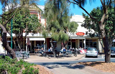 Outdoor cafe with tables and people, surrounded by trees, under a clear blue sky.