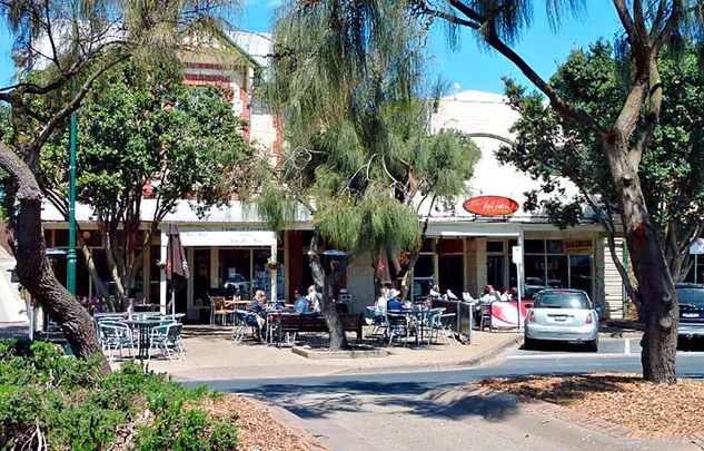 Outdoor cafe with tables and people, surrounded by trees, under a clear blue sky.