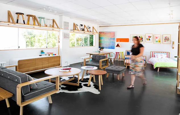 Contemporary furniture showroom with wooden chairs, colorful stools, and artwork on the walls. Woman walking through.